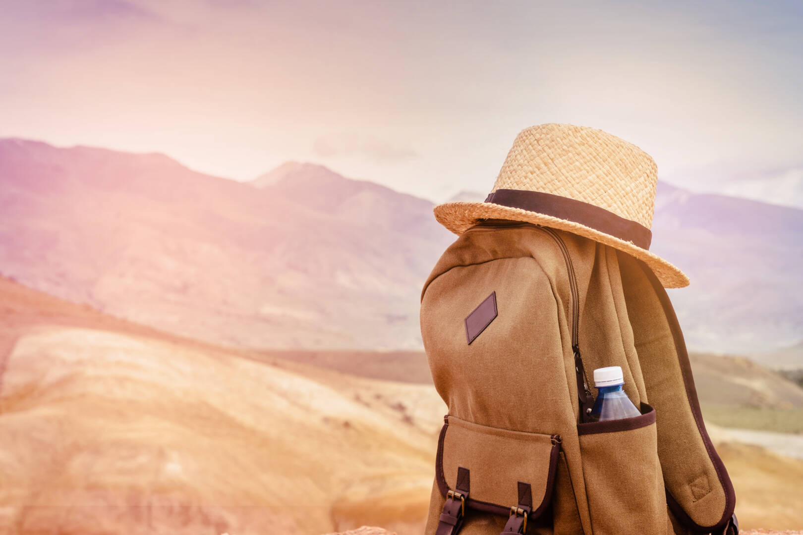 Beige backpack, bottle of water and straw hat on on top of the mountain. Active travel concept. Toning sunrise.